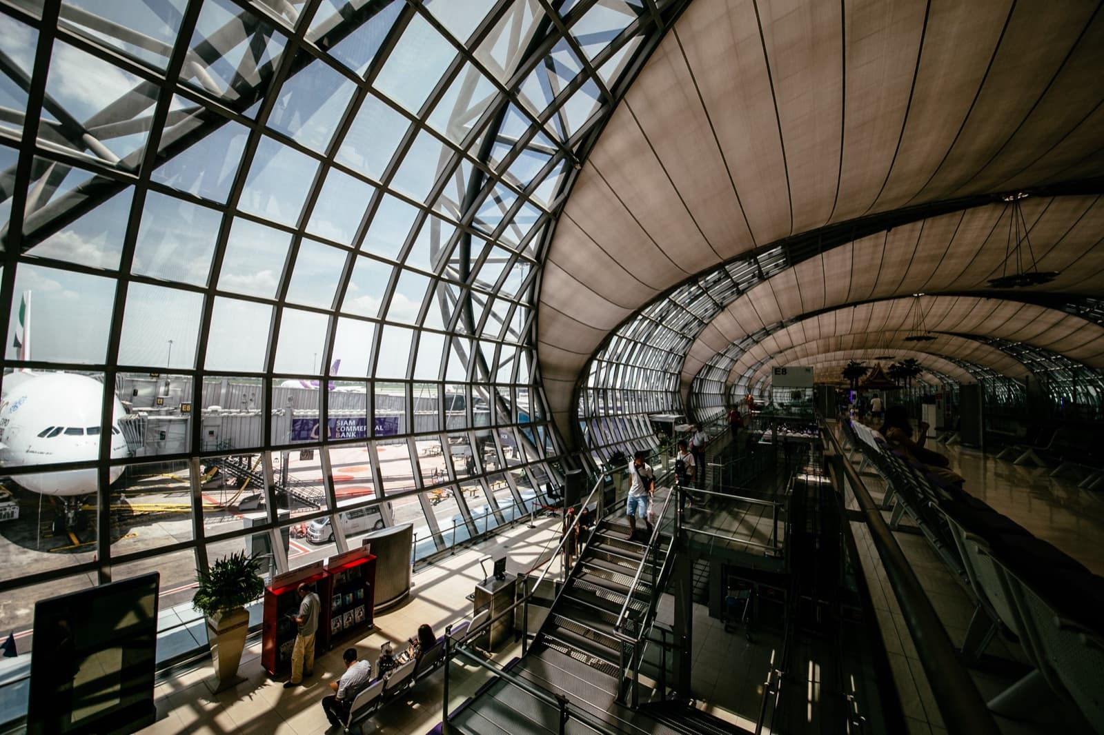 A sunlit glass-roofed airport terminal with an aircraft at the gate