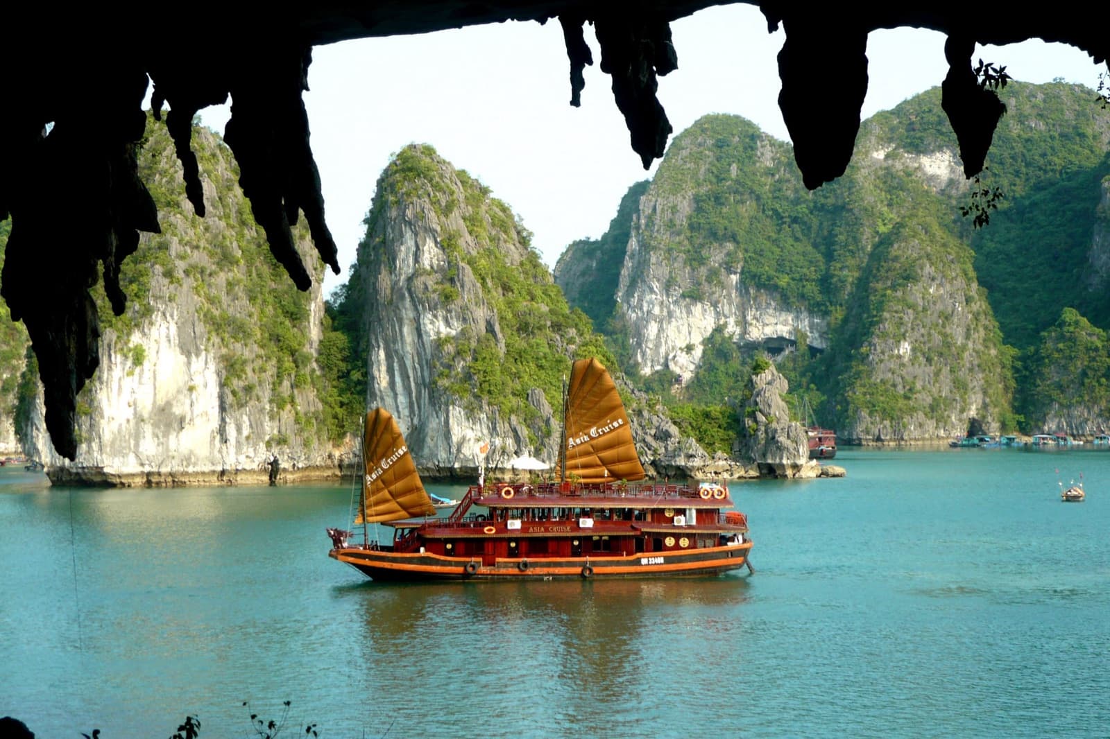 Ha Long Bay from a cave mouth: a traditional sailing cruise junk on turquoise water among green limestone islands