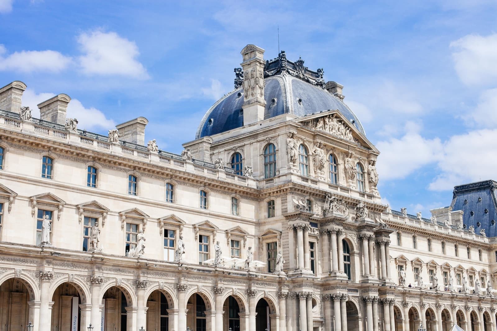 Cung điện Louvre ở Paris: mặt tiền đá màu kem chạm khắc tinh xảo và mái vòm xám xanh dưới bầu trời xanh