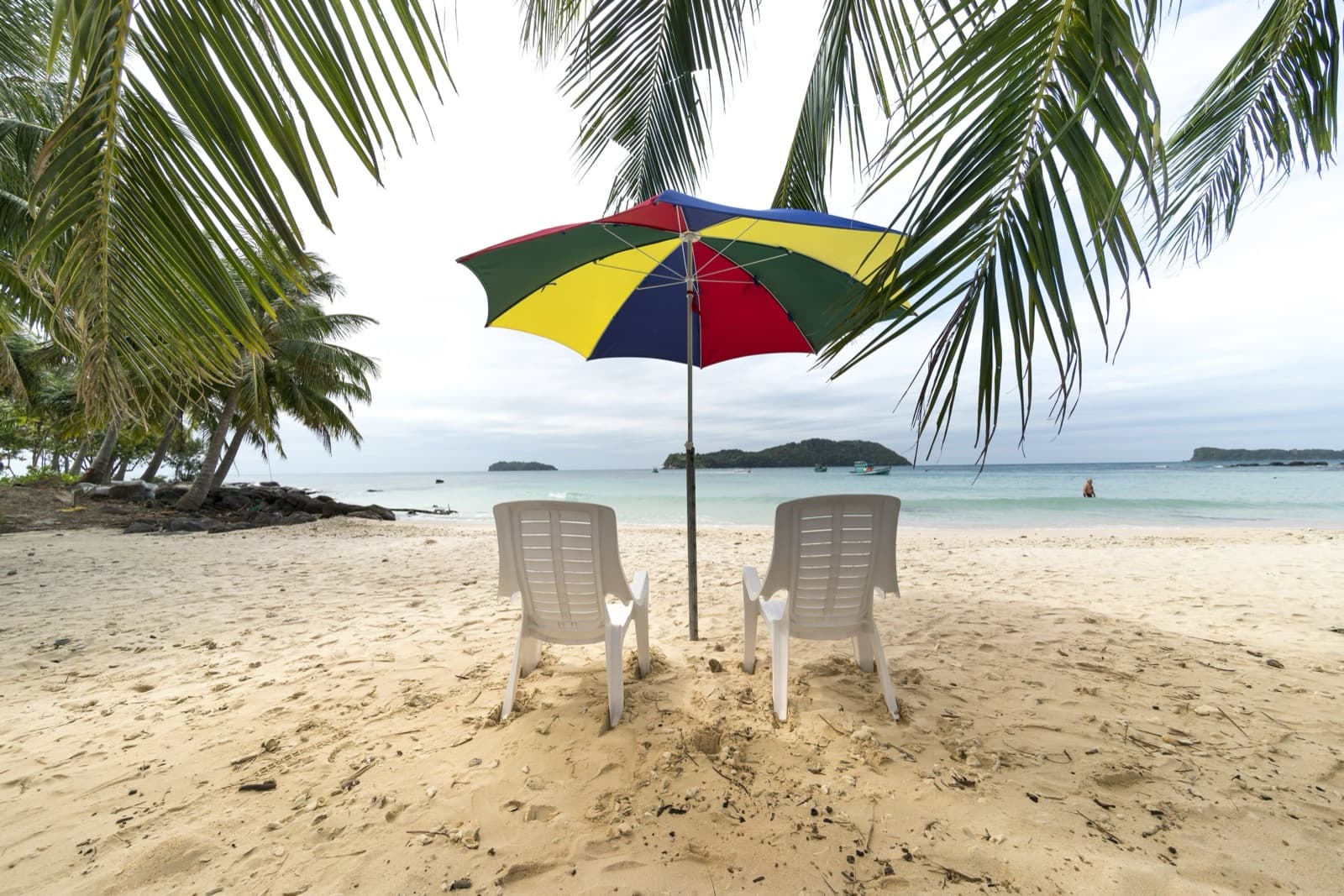 A Phu Quoc beach with two white loungers under a colourful umbrella and palm fronds, turquoise sea and small islands beyond